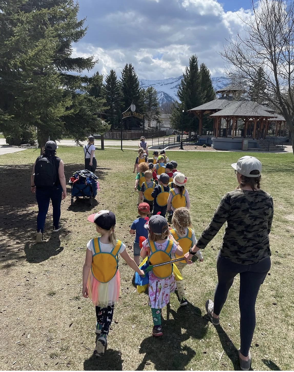 Little Me children in yellow vests walking through a Fernie park with the mountains behind them