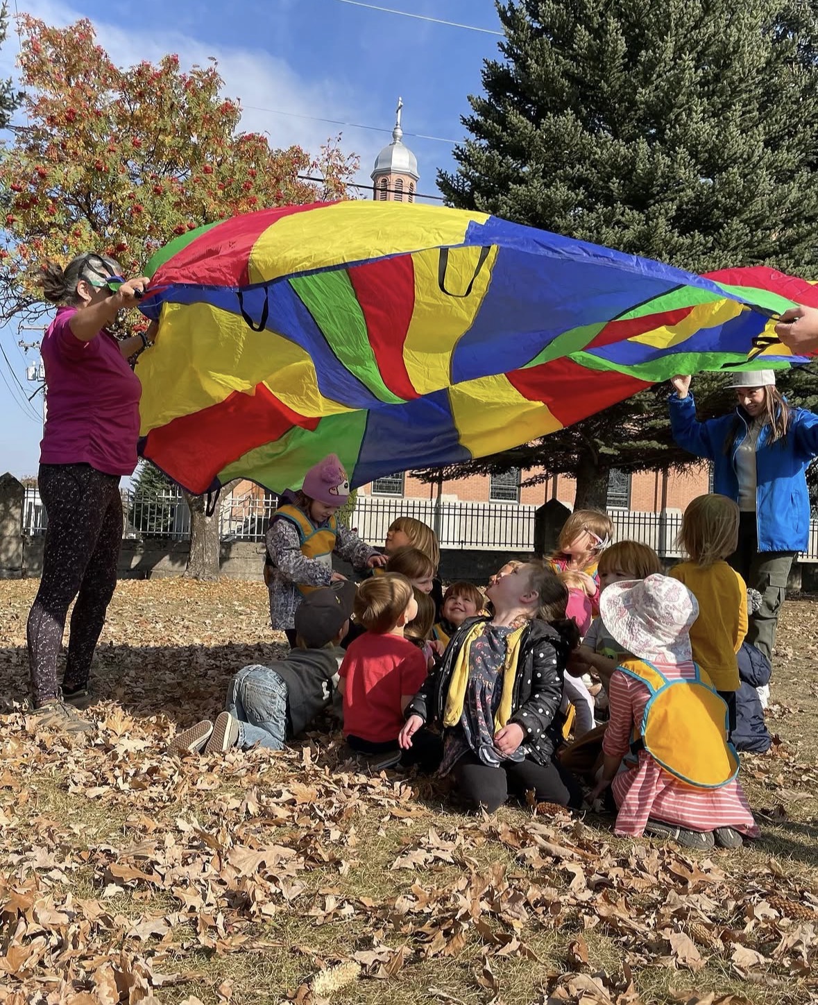 Little Me children laughing and playing together under a colourful parachute outdoors